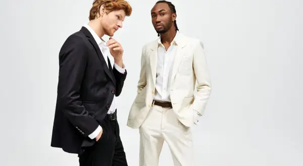 Due uomini in abiti eleganti. Uno con giacca nera e pantaloni neri, l'altro con giacca e pantaloni color crema. Il **slim fit significato** è evidente nella vestibilità moderna.
