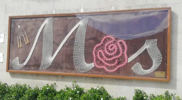 Letras de encaje blanco y rosa forman "Mos" en un letrero en el Pazo de Mos, con herramientas de encaje a la izquierda.