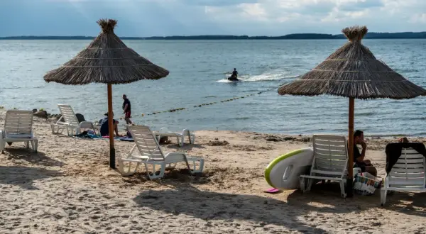 Słoneczne jezioro z plażą, leżakami i parasolami. Na wodzie widać skuter wodny.