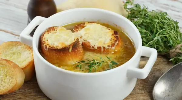 Aksamitna zupa cebulowa przepis, podana w białej miseczce, z grzankami zapieczonymi z serem i gałązką tymianku. Obok kromki chleba.
