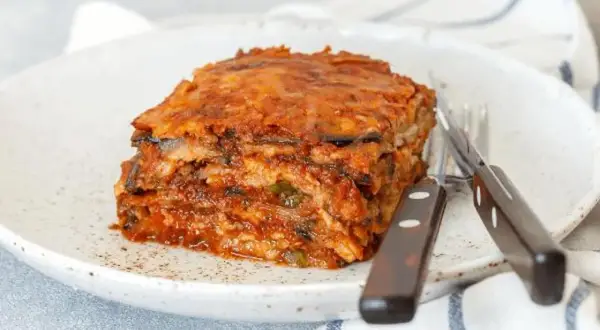 Porción de pastel de berenjenas, con capas de berenjena y salsa de tomate, servido en un plato blanco con cubiertos. ¡Un pastel de berenjenas facilisimo!