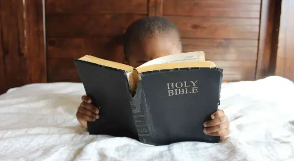Dziecko z zaciekawieniem czyta Biblię, leżąc w łóżku. Słowa "Holy Bible" są widoczne na okładce.