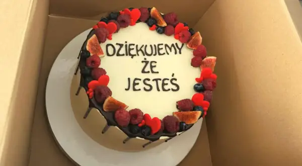 Okrągły tort z białym kremem, ozdobiony owocami i serduszkami. Krótki napis na tort urodzinowy: "DZIĘKUJEMY ŻE JESTEŚ".