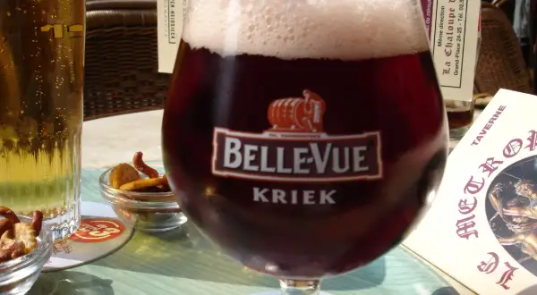 Szklanka piwa cherry Kriek Bellevue z pianką, obok jasne piwo i miseczki z przekąskami.