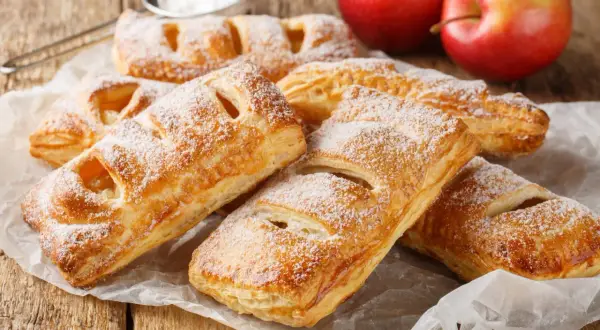 Szybkie ciasto francuskie z jabłkami, posypane cukrem pudrem, leży na papierze. Obok dwa czerwone jabłka.