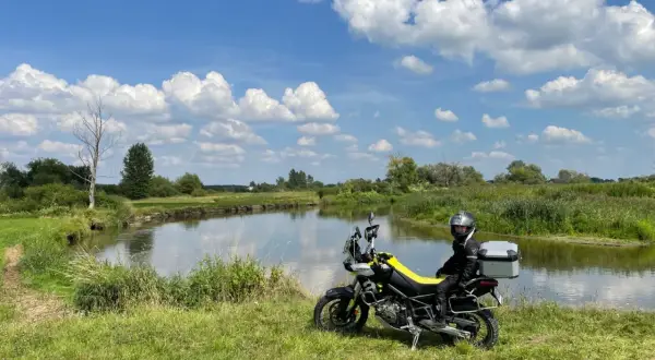 Motocyklista w kasku siedzi na motocyklu obok rzeki, na tle zielonej łąki i błękitnego nieba z chmurami.