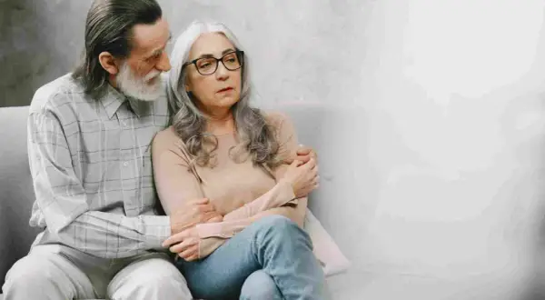 Coppia anziana seduta sul divano. Lui la conforta, forse parlando di come la pensione di invalidità è reversibile.