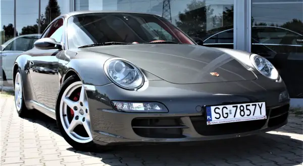 Szary Porsche 997 z czerwonym wnętrzem, zaparkowany na kostce brukowej przed budynkiem z lustrzaną fasadą.