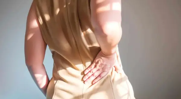 Une femme se tient le bas du dos, ressentant une lourdeur. La lumière du soleil projette des ombres sur sa peau.