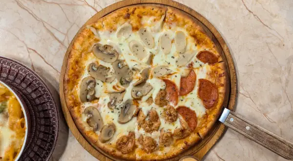 Pizza quattro stagioni avec champignons, saucisses, pepperoni et poulet frit. Une part de pâtes gratinées est visible à gauche.