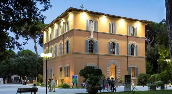 Edificio giallo illuminato nel parco della Versiliana, con persone che passeggiano e un'opera d'arte esposta.