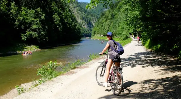 Dziewczyna na rowerze na malowniczej trasie rowerowej w Szczawnicy. W tle Dunajec z tratwami i zalesione góry.