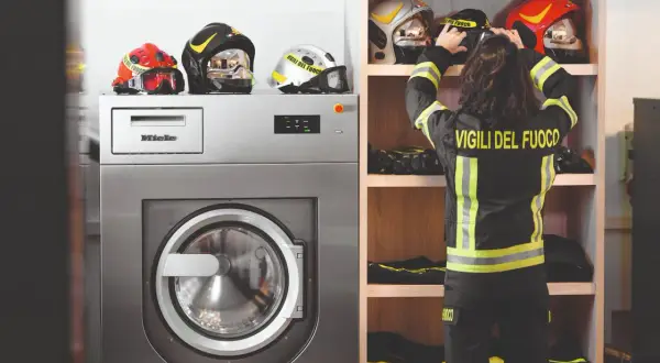 Vigile del Fuoco sistema i caschi accanto a una lavatrice industriale, pronta per il lavaggio abiti da lavoro.