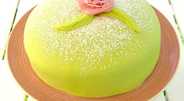 En vacker prinsesstårta med grön marsipan, pudrad med florsocker och dekorerad med en rosa ros. Perfekt för ett prinsesstårta recept!
