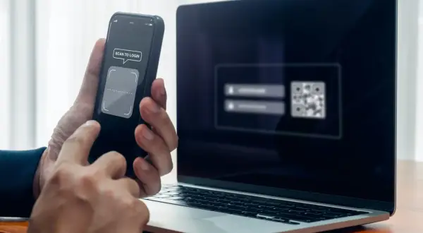 Ręce trzymają telefon z napisem "SCAN TO LOGIN", obok laptop z kodem QR. Szybkie logowanie, jak zeskanować kod na telefon.