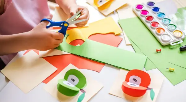 Manos pequeñas cortando papel para manualidades para niños de 3 a 5 años. Pinturas, tijeras y frutas de papel decoran la mesa.