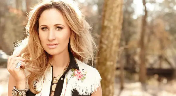 Jessi Alexander, a blonde woman with striking eyes, poses outdoors in a fringed vest adorned with floral embroidery.