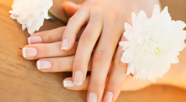 Delikatne dłonie z francuskim manicure, otoczone białymi kwiatami. Paznokcie wyglądają zdrowo, nie są suche.