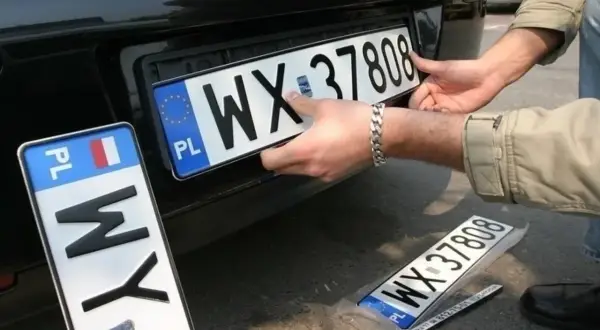 Montaż nowych tablic rejestracyjnych. Jedna z nich, z numerem WX 37808, jest właśnie wkładana. Obok leży kolejna, zapakowana.