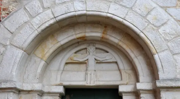 Kamienna płaskorzeźba postaci z koroną na głowie, rozłożonymi ramionami i ozdobnym pasem, osadzona w łuku portalu. To przykład sztuki romańskiej.