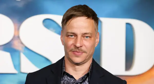 Tom Wlaschiha, mit einem Schnurrbart und blauen Augen, posiert vor einem Hintergrund mit großen weißen Buchstaben. Er denkt vielleicht an seine tom wlaschiha kinder.