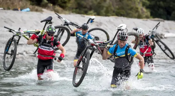 Teilnehmer eines **adventure racing** durchqueren mit Fahrrädern auf den Schultern einen Fluss.