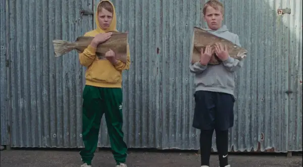 Två barn, som påminner om Sverrir Gudnason som barn, håller varsin fisk framför en plåtvägg.