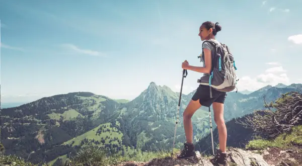 Młoda kobieta z plecakiem i kijkami trekkingowymi podziwia panoramę gór. Idealny widok dla tych, którzy zastanawiają się, jak zacząć chodzić po górach.