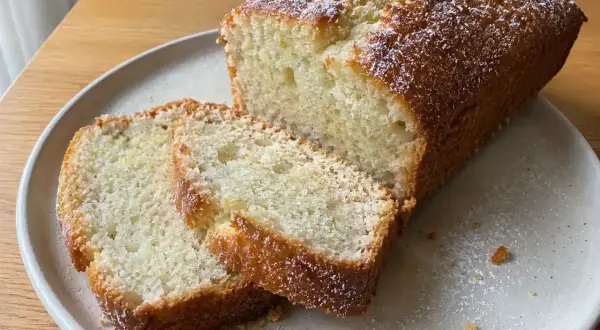 En saftig vegansk sockerkaka, pudrad med florsocker, ligger på en tallrik. Två skivor är upplagda bredvid.