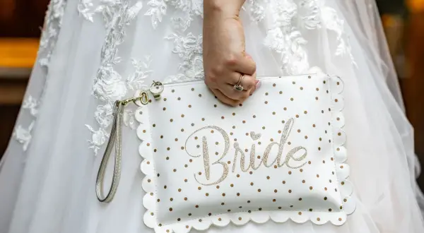 Biała kopertówka w złote kropki z napisem "Bride" to niezbędnik panny młodej. Dopełnia suknię ślubną.