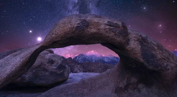 A natural rock arch frames a majestic mountain range under a breathtaking starry sky.