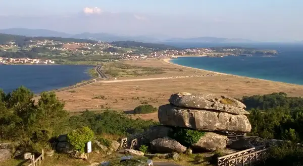 Vista panorámica desde el mirador de Siradella. Rocas gigantescas y un puente de madera en primer plano, con un paisaje de costa, mar azul y un pueblo al fondo.