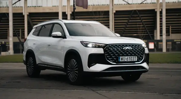 Biały Chery Tiggo 8 z charakterystycznym grillem i światłami LED, zaparkowany przed trybunami stadionu.