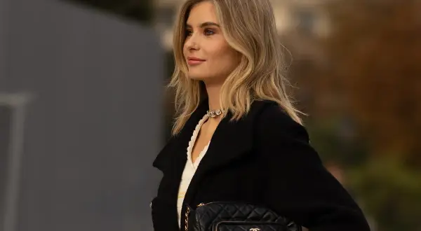 Kobieta z beżowym blondem, w czarnym płaszczu i z pikowaną torebką Chanel, prezentuje elegancką stylizację.