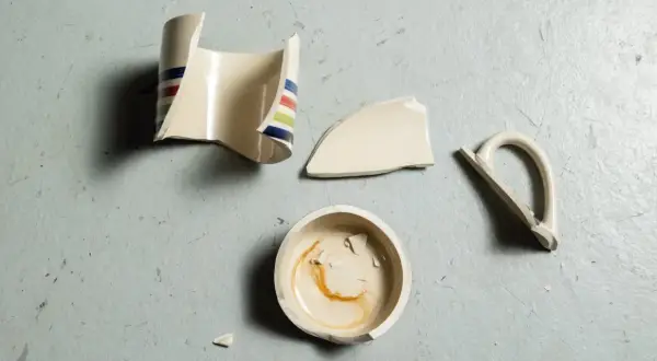 Rozbite fragmenty ceramicznego kubka z kolorowymi paskami leżą na szarym tle. Widać podstawę, korpus, uchwyt i kilka mniejszych kawałków. To materiał do nauki, jak skleić kubek ceramiczny.