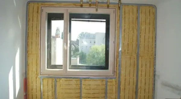 Izolacja akustyczna wokół okna, z widocznym materiałem w kolorze piaskowym na ścianie, przygotowana do dalszych prac wykończeniowych.