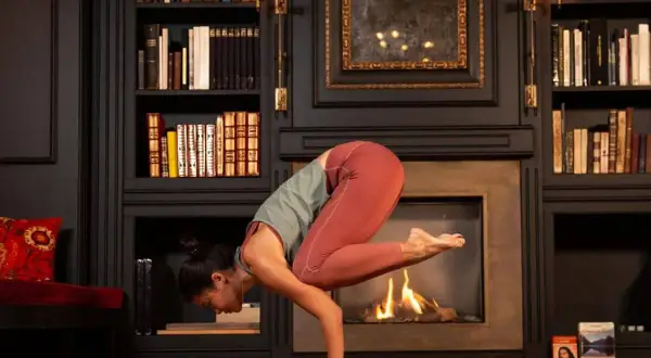 Mujer en postura de grulla, inspirada en frases yoga Patanjali. Chimenea encendida y estanterías llenas de libros.