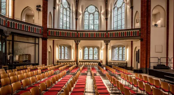 Innenraum einer epo church mit vielen roten Stuhlreihen und großen Bogenfenstern. Ein Mann sitzt allein.