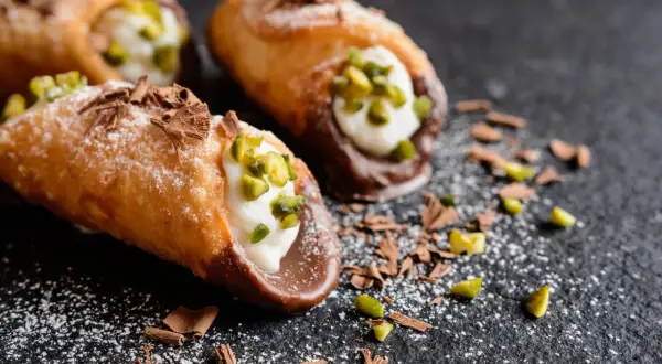 Trois cannoli salés, garniture crémeuse, pistaches concassées et copeaux de chocolat, saupoudrés de sucre glace sur fond sombre.
