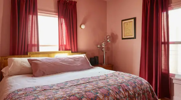 A bedroom with a large bed covered by a colorful quilt. Red curtains hang beside the bed, effectively creating a cozy nook and demonstrating how to hide a window behind a bed.