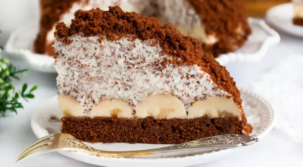 Kawałek czekoladowego ciasta z bananami i kremem kokosowym, posypany okruszkami.
