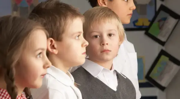 Cztery dzieci w szkolnych strojach, skupione na czymś poza kadrem. W tle widać prace plastyczne, sugerujące naukę i nauczanie indywidualne.