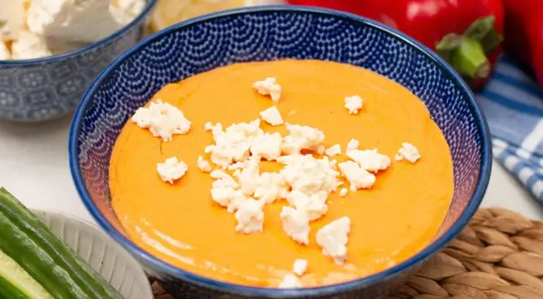 Cremiger Paprika-Feta-Dip in blauer Schüssel, garniert mit Feta-Käse. Dazu Gurkensticks und rote Paprika.