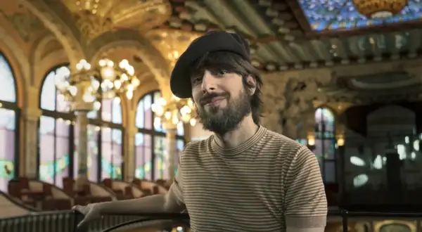 David Ucles sonríe con un gorro y una camiseta a rayas, en un salón con vidrieras y lámparas doradas.