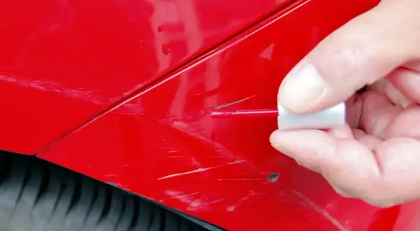 Dłoń z pędzelkiem aplikuje lakier na porysowane auto.