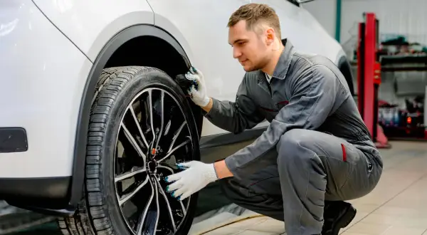 Mécanicien en combinaison grise, avec des gants, examine la roue d'une voiture blanche. Il semble chercher comment dégripper un étrier de frein sans démonter.