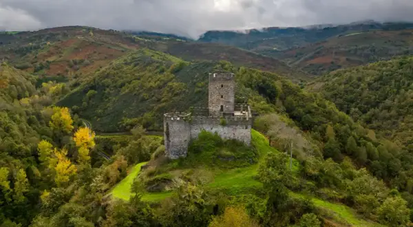 El **castillo de Doiras** se alza majestuoso sobre una colina verde, rodeado de montañas boscosas y un cielo nublado.
