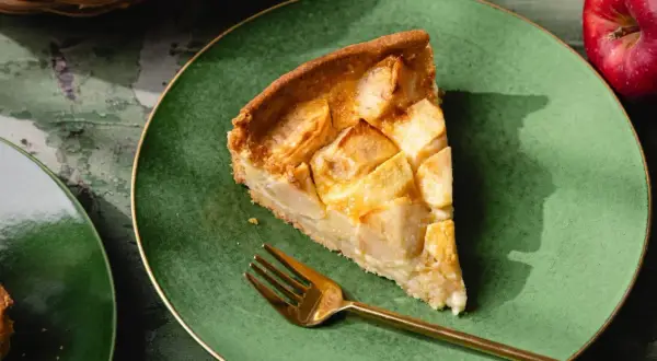Une part de tarte normande aux pommes, dorée et appétissante, sur une assiette verte avec une fourchette dorée.