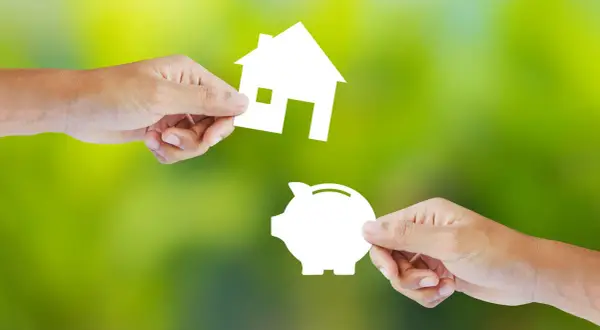 Manos intercambian una casa y una alcancía, simbolizando la decisión de alquilar o comprar.