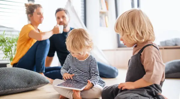 Rozpieszczone dziecko pochłonięte tabletem, podczas gdy rodzice w tle piją kawę.